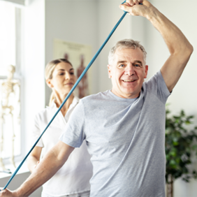 Ein älterer Mann in einem grauen T-Shirt führt eine Dehnübung mit einem blauen Widerstandsband durch. Eine Frau, möglicherweise eine Physiotherapeutin, steht hinter ihm und unterstützt ihn bei der Übung. Im Hintergrund ist ein helles Zimmer mit einem großen Fenster zu sehen, durch das Tageslicht hereinfällt.
