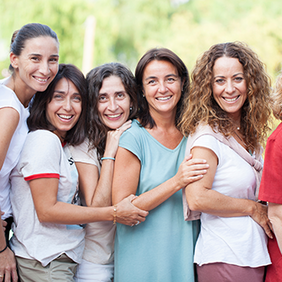 Auf dem Bild stehen sieben Frauen nebeneinander. Alle haben unterschiedlichen Frisuren und unterschiedliche Kleidnug an. Alle lachen und sind fröhlich und gucken nach vorne.