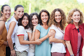 Auf dem Bild stehen sieben Frauen nebeneinander. Alle haben unterschiedlichen Frisuren und unterschiedliche Kleidnug an. Alle lachen und sind fröhlich und gucken nach vorne.