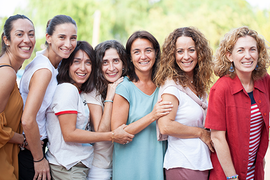 Auf dem Bild stehen sieben Frauen nebeneinander. Alle haben unterschiedlichen Frisuren und unterschiedliche Kleidnug an. Alle lachen und sind fröhlich und gucken nach vorne.