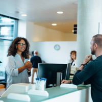 In einem modernen, hellen Bürogebäude stehen drei Personen an einem Empfangstresen. Eine Frau mit lockigem Haar und Brille steht hinter dem Tresen und lächelt freundlich. Vor dem Tresen stehen ein Mann und eine Frau, die beide ebenfalls lächeln. Im Hintergrund sind weitere Personen zu sehen, die sich in der offenen und gut beleuchteten Umgebung bewegen. Pflanzen und moderne Möbel tragen zur angenehmen Atmosphäre bei.