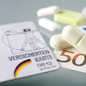 Auf dem Bild sind eine Krankenversicherungskarte, mehrere Euro-Banknoten und -Münzen sowie einige Tabletten zu sehen. Die Krankenversicherungskarte liegt links im Vordergrund, während die Banknoten und Münzen rechts daneben platziert sind. Die Tabletten befinden sich in der Mitte des Bildes.
