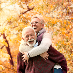 Ein älteres Paar genießt einen sonnigen Herbsttag im Freien. Die Frau, die eine Brille trägt, umarmt den Mann von hinten und beide lachen herzlich. Sie sind von herbstlich gefärbten Bäumen umgeben, deren Blätter in warmen Gelb- und Orangetönen leuchten. Der Mann trägt einen weinroten Pullover, während die Frau eine helle Jacke anhat.