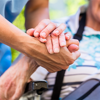 Auf dem Bild sieht man eine Nahaufnahme von zwei Händen, die sich halten. Eine Hand gehört zu einer älteren Person, die in einem Rollstuhl sitzt und ein gestreiftes Hemd trägt. Die andere Hand gehört zu einer jüngeren Person, die Pflege leistet. Im Hintergrund ist das Gesicht der älteren Person leicht unscharf zu erkennen, sie scheint zu lächeln.