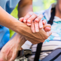 Auf dem Bild sieht man eine Nahaufnahme von zwei Händen, die sich halten. Eine Hand gehört zu einer älteren Person, die in einem Rollstuhl sitzt und ein gestreiftes Hemd trägt. Die andere Hand gehört zu einer jüngeren Person, die Pflege leistet. Im Hintergrund ist das Gesicht der älteren Person leicht unscharf zu erkennen, sie scheint zu lächeln.