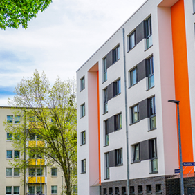 Das Bild zeigt eine städtische Wohngegend mit modernen Mehrfamilienhäusern. Die Gebäude haben eine helle Fassade, wobei eines der Häuser durch orangefarbene Akzente hervorsticht. Die Fenster sind gleichmäßig angeordnet und einige Balkone sind zu sehen. 
