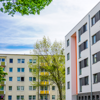 Das Bild zeigt eine städtische Wohngegend mit modernen Mehrfamilienhäusern. Die Gebäude haben eine helle Fassade, wobei eines der Häuser durch orangefarbene Akzente hervorsticht. Die Fenster sind gleichmäßig angeordnet und einige Balkone sind zu sehen. 
