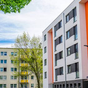 Das Bild zeigt eine städtische Wohngegend mit modernen Mehrfamilienhäusern. Die Gebäude haben eine helle Fassade, wobei eines der Häuser durch orangefarbene Akzente hervorsticht. Die Fenster sind gleichmäßig angeordnet und einige Balkone sind zu sehen. 