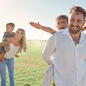 Auf dem Bild ist eine Familie zu sehen, die auf einer Wiese sind. Im Hintergrund trägt die Frau ein Kind auf dem Rücken. Der Mann im Vordergrund trägt auch ein Kind auf dem Rücken. Das Kind im Vordergrund streckt die Arme aus. 