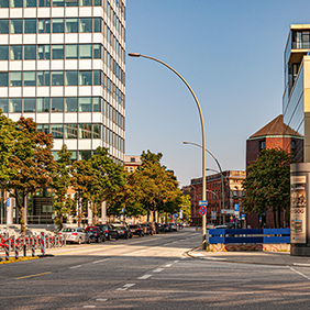 Eine breite, mehrspurige Straße verläuft durch ein urbanes Gebiet mit modernen Bürogebäuden und Bäumen am Straßenrand. Links ist ein Eingang zur U-Bahn-Station erkennbar. Mehrere geparkte Autos stehen am Straßenrand, daneben befinden sich Fahrradständer mit Leihfahrrädern. Rechts ragt ein rotes Backsteingebäude mit Glasfassade in das Bild. Die Straße ist fast leer, der Himmel ist blau und klar.