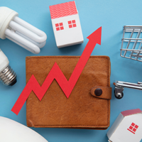 Das Bild zeigt eine braune Geldbörse, auf der ein roter Pfeil in Form eines steigenden Diagramms platziert ist. Um die Geldbörse herum sind verschiedene Gegenstände angeordnet: zwei Glühbirnen, ein Miniatur-Einkaufswagen und zwei kleine Häuser. Der Hintergrund ist hellblau. Das Bild symbolisiert steigende Kosten oder Preise in verschiedenen Lebensbereichen wie Energie, Wohnen und Konsumgüter.