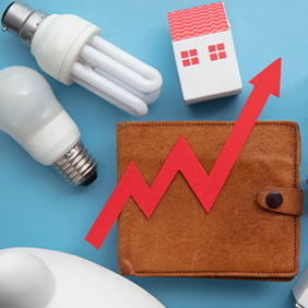 Das Bild zeigt eine braune Geldbörse, auf der ein roter Pfeil in Form eines steigenden Diagramms platziert ist. Um die Geldbörse herum sind verschiedene Gegenstände angeordnet: zwei Glühbirnen, ein Miniatur-Einkaufswagen und zwei kleine Häuser. Der Hintergrund ist hellblau. Das Bild symbolisiert steigende Kosten oder Preise in verschiedenen Lebensbereichen wie Energie, Wohnen und Konsumgüter.