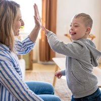 Ein kleiner Junge und eine Frau sitzen sich in einem gemütlichen Wohnzimmer gegenüber. Der Junge trägt einen grauen Kapuzenpullover und gibt der Frau, die ein gestreiftes Hemd trägt, ein High-Five. 