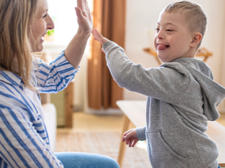Ein kleiner Junge und eine Frau sitzen sich in einem gemütlichen Wohnzimmer gegenüber. Der Junge trägt einen grauen Kapuzenpullover und gibt der Frau, die ein gestreiftes Hemd trägt, ein High-Five. 
