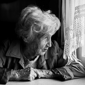 Eine ältere Frau mit welligem, grauem Haar sitzt an einem Tisch und schaut nachdenklich aus einem Fenster. Sie trägt eine Jacke und hat ihre Hände vor sich auf dem Tisch gefaltet. Das Licht, das durch das Fenster fällt, beleuchtet ihr Gesicht. Die Szene ist in Schwarz-Weiß gehalten.