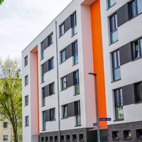 Das Bild zeigt eine städtische Wohngegend mit modernen Mehrfamilienhäusern. Die Gebäude haben eine helle Fassade, wobei eines der Häuser durch orangefarbene Akzente hervorsticht. Die Fenster sind gleichmäßig angeordnet und einige Balkone sind zu sehen. 