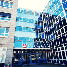 Das Bild zeigt die Fassade eines modernen Bürogebäudes mit großen Glasfenstern. Das Gebäude hat mehrere Stockwerke und eine Kombination aus blauen Glasflächen und beigen Steinwänden. Über dem Eingang befindet sich ein Schild mit der Aufschrift "Agentur für Arbeit" und einem roten Logo. Der Eingangsbereich ist durch eine Glasüberdachung geschützt, und es gibt mehrere Türen, die in das Gebäude führen.
