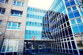 Das Bild zeigt die Fassade eines modernen Bürogebäudes mit großen Glasfenstern. Das Gebäude hat mehrere Stockwerke und eine Kombination aus blauen Glasflächen und beigen Steinwänden. Über dem Eingang befindet sich ein Schild mit der Aufschrift "Agentur für Arbeit" und einem roten Logo. Der Eingangsbereich ist durch eine Glasüberdachung geschützt, und es gibt mehrere Türen, die in das Gebäude führen.