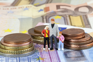 Das Bild zeigt drei Miniaturfiguren, die eine Familie darstellen, bestehend aus einem Erwachsenen und zwei Kindern. Sie stehen auf einem Haufen von Euro-Münzen und -Scheinen. Die Münzen sind in verschiedenen Größen gestapelt, und die Scheine sind im Hintergrund sichtbar. 