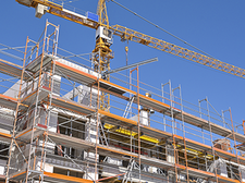 Das Bild zeigt eine Baustelle, auf der ein mehrstöckiges Gebäude errichtet wird. Das Gebäude ist von einem Gerüst umgeben, das den Arbeitern Zugang zu verschiedenen Ebenen ermöglicht. Im Hintergrund ragt ein großer Baukran in den klaren blauen Himmel, der für den Transport von Baumaterialien verwendet wird. 