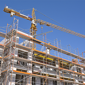 Das Bild zeigt eine Baustelle, auf der ein mehrstöckiges Gebäude errichtet wird. Das Gebäude ist von einem Gerüst umgeben, das den Arbeitern Zugang zu verschiedenen Ebenen ermöglicht. Im Hintergrund ragt ein großer Baukran in den klaren blauen Himmel, der für den Transport von Baumaterialien verwendet wird.