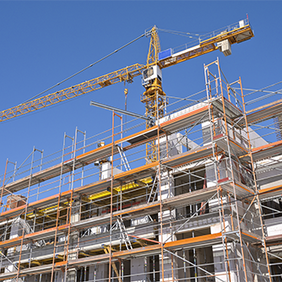 Das Bild zeigt eine Baustelle, auf der ein mehrstöckiges Gebäude errichtet wird. Das Gebäude ist von einem Gerüst umgeben, das den Arbeitern Zugang zu verschiedenen Ebenen ermöglicht. Im Hintergrund ragt ein großer Baukran in den klaren blauen Himmel, der für den Transport von Baumaterialien verwendet wird.