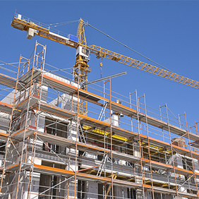 Das Bild zeigt eine Baustelle, auf der ein mehrstöckiges Gebäude errichtet wird. Das Gebäude ist von einem Gerüst umgeben, das den Arbeitern Zugang zu verschiedenen Ebenen ermöglicht. Im Hintergrund ragt ein großer Baukran in den klaren blauen Himmel, der für den Transport von Baumaterialien verwendet wird.