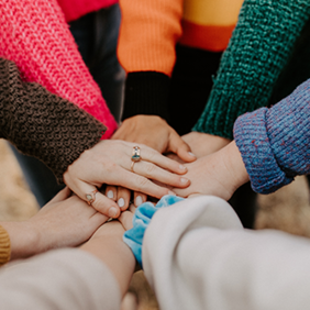Eine Gruppe von Menschen steht im Kreis und legt ihre Hände übereinander in die Mitte. Sie tragen bunte Pullover in verschiedenen Farben wie Pink, Orange, Grün und Blau.