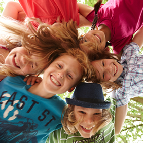 Auf dem Bild sind sechs fröhliche Kinder zu sehen, die in einem Kreis stehen und ihre Köpfe zusammenstecken. Sie lachen und schauen nach unten in die Kamera. Die Kinder tragen bunte Kleidung. Im Hintergrund sind grüne Blätter von Bäumen zu erkennen.