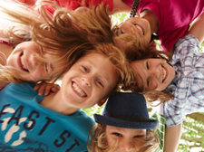 Auf dem Bild sind sechs fröhliche Kinder zu sehen, die in einem Kreis stehen und ihre Köpfe zusammenstecken. Sie lachen und schauen nach unten in die Kamera. Die Kinder tragen bunte Kleidung. Im Hintergrund sind grüne Blätter von Bäumen zu erkennen.