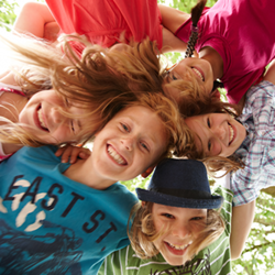 Auf dem Bild sind sechs fröhliche Kinder zu sehen, die in einem Kreis stehen und ihre Köpfe zusammenstecken. Sie lachen und schauen nach unten in die Kamera. Die Kinder tragen bunte Kleidung. Im Hintergrund sind grüne Blätter von Bäumen zu erkennen.