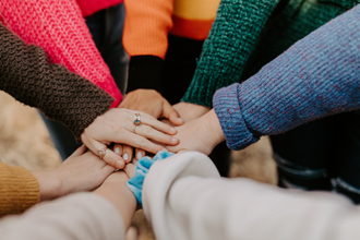Eine Gruppe von Menschen steht im Kreis und legt ihre Hände übereinander in die Mitte. Sie tragen bunte Pullover in verschiedenen Farben wie Pink, Orange, Grün und Blau.
