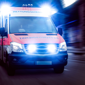 Ein Rettungswagen fährt mit eingeschaltetem Blaulicht und Sirene. Das Fahrzeug ist in den Farben Rot und Weiß gehalten und trägt die Aufschrift Rettungsdienst auf der Front. Die Scheinwerfer und das Blaulicht leuchten hell.
