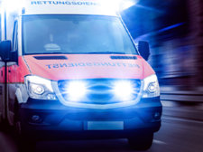Ein Rettungswagen fährt mit eingeschaltetem Blaulicht und Sirene. Das Fahrzeug ist in den Farben Rot und Weiß gehalten und trägt die Aufschrift Rettungsdienst auf der Front. Die Scheinwerfer und das Blaulicht leuchten hell.
