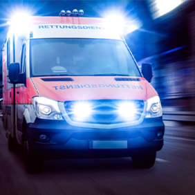 Ein Rettungswagen fährt mit eingeschaltetem Blaulicht und Sirene. Das Fahrzeug ist in den Farben Rot und Weiß gehalten und trägt die Aufschrift Rettungsdienst auf der Front. Die Scheinwerfer und das Blaulicht leuchten hell.