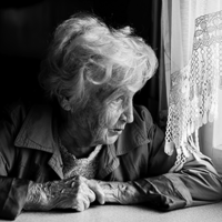Eine ältere Frau mit welligem, grauem Haar sitzt an einem Tisch und schaut nachdenklich aus einem Fenster. Sie trägt eine Jacke und hat ihre Hände vor sich auf dem Tisch gefaltet. Das Licht, das durch das Fenster fällt, beleuchtet ihr Gesicht. Die Szene ist in Schwarz-Weiß gehalten.