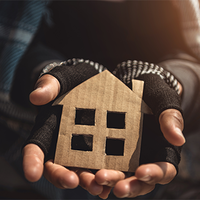 Eine Person hält ein kleines, aus Pappe ausgeschnittenes Symbol in Form eines Hauses in ihren Händen. Die Hände tragen fingerlose Handschuhe, die auf Kälte und möglicherweise Wohnungslosigkeit hinweisen. Die Kleidung wirkt abgenutzt. Das Licht fällt warm von der Seite ein und beleuchtet das Haus-Symbol.