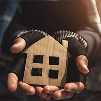 Eine Person hält ein kleines, aus Pappe ausgeschnittenes Symbol in Form eines Hauses in ihren Händen. Die Hände tragen fingerlose Handschuhe, die auf Kälte und möglicherweise Wohnungslosigkeit hinweisen. Die Kleidung wirkt abgenutzt. Das Licht fällt warm von der Seite ein und beleuchtet das Haus-Symbol.