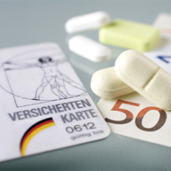 Auf dem Bild sind eine Krankenversicherungskarte, mehrere Euro-Banknoten und -Münzen sowie einige Tabletten zu sehen. Die Krankenversicherungskarte liegt links im Vordergrund, während die Banknoten und Münzen rechts daneben platziert sind. Die Tabletten befinden sich in der Mitte des Bildes.