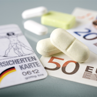 Auf dem Bild sind eine Krankenversicherungskarte, mehrere Euro-Banknoten und -Münzen sowie einige Tabletten zu sehen. Die Krankenversicherungskarte liegt links im Vordergrund, während die Banknoten und Münzen rechts daneben platziert sind. Die Tabletten befinden sich in der Mitte des Bildes.