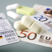Auf dem Bild sind eine Krankenversicherungskarte, mehrere Euro-Banknoten und -Münzen sowie einige Tabletten zu sehen. Die Krankenversicherungskarte liegt links im Vordergrund, während die Banknoten und Münzen rechts daneben platziert sind. Die Tabletten befinden sich in der Mitte des Bildes.