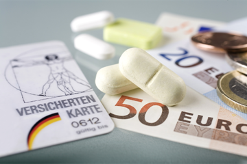 Auf dem Bild sind eine Krankenversicherungskarte, mehrere Euro-Banknoten und -Münzen sowie einige Tabletten zu sehen. Die Krankenversicherungskarte liegt links im Vordergrund, während die Banknoten und Münzen rechts daneben platziert sind. Die Tabletten befinden sich in der Mitte des Bildes.