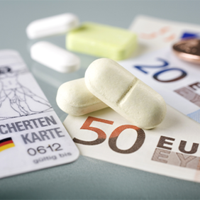 Auf dem Bild sind eine Krankenversicherungskarte, mehrere Euro-Banknoten und -Münzen sowie einige Tabletten zu sehen. Die Krankenversicherungskarte liegt links im Vordergrund, während die Banknoten und Münzen rechts daneben platziert sind. Die Tabletten befinden sich in der Mitte des Bildes.