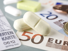 Auf dem Bild sind eine Krankenversicherungskarte, mehrere Euro-Banknoten und -Münzen sowie einige Tabletten zu sehen. Die Krankenversicherungskarte liegt links im Vordergrund, während die Banknoten und Münzen rechts daneben platziert sind. Die Tabletten befinden sich in der Mitte des Bildes.
