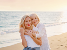 Das Bild zeigt zwei Frauen, die sich an einem Strand umarmen. Die jüngere Frau steht vorne und trägt ein weißes T-Shirt und eine blaue Jeans. Die ältere Frau steht hinter ihr und hat ihre Arme um die Schultern der jüngeren Frau gelegt. Beide lächeln glücklich in die Kamera. Im Hintergrund ist das Meer zu sehen.