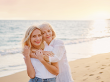Das Bild zeigt zwei Frauen, die sich an einem Strand umarmen. Die jüngere Frau steht vorne und trägt ein weißes T-Shirt und eine blaue Jeans. Die ältere Frau steht hinter ihr und hat ihre Arme um die Schultern der jüngeren Frau gelegt. Beide lächeln glücklich in die Kamera. Im Hintergrund ist das Meer zu sehen.