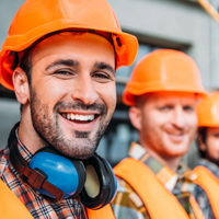 Das Bild zeigt drei Bauarbeiter, die Schutzhelme und reflektierende Westen tragen. Der Arbeiter im Vordergrund lächelt in die Kamera und trägt zusätzlich Gehörschutz um den Hals. Die beiden anderen Arbeiter im Hintergrund sind ebenfalls in Arbeitskleidung. Im Hintergrund ist eine Baustelle zu erkennen.