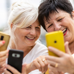 Drei ältere Frauen sitzen zusammen und lachen, während sie auf ihre Smartphones schauen. Alle drei halten ihre Telefone in den Händen und wirken entspannt und glücklich. Die Frau links trägt eine Brille und ein hellblaues Oberteil, die Frau in der Mitte hat weißes Haar und trägt ein weißes Oberteil, und die Frau rechts hat dunkles Haar und trägt ein weißes Oberteil.