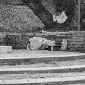 Schwarz-Weiß: Das Bild zeigt eine Szene in einem Park, in der eine Person auf einer Bank unter einer Decke schläft. Die Bank befindet sich vor einer niedrigen Backsteinmauer, hinter der ein großer Baum steht. Auf der Mauer liegt eine weitere Decke oder ein Tuch. Es gibt einige Stufen, die zu der Bank führen. Im Hintergrund sind kahle Bäume und Laub zu sehen, was auf die Herbst- oder Winterzeit hindeutet.