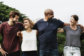 Eine Gruppe von vier Personen steht im Freien auf einer Wiese. Sie haben die Arme umeinander gelegt und lächeln. Im Hintergrund sind Bäume zu sehen, und der Himmel ist klar. Die Menschen tragen legere Kleidung.
